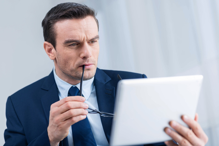Market situation. Concentrated budding attractive man bitting his glasses while holding tablet and looking at itの写真素材