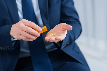 Health important. Close up of  strong man hands where yellow pills lying on the palm while other hand holding bottle and man wearing suitの写真素材