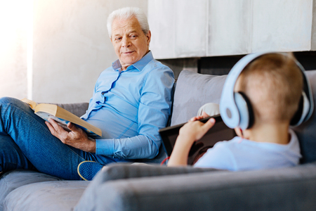 Family evening. Happy nice cheerful man sitting together with his grandson and looking at his while reading a bookの写真素材