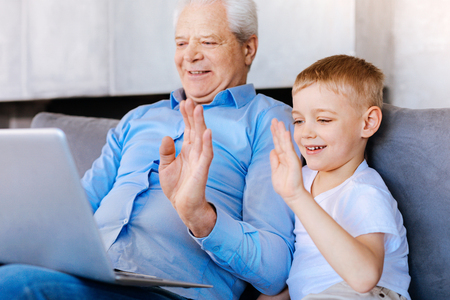 Online call. Joyful nice pleasant boy and his grandfather looking at the laptop screen and waving their hands while making an internet callの写真素材