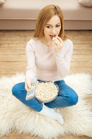 Great weekend. Good-looking cheerful young blond dark-eyed woman holding a remote control and eating...