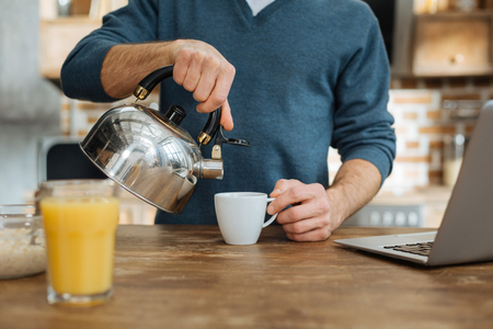 Making tea. Delighted man standing near table and raising right hand while pouring down waterの写真素材
