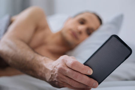 Turn it off. Low-spirited man taking his gadget while looking at clock and being in bedの写真素材