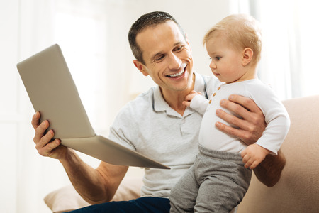 Funny cartoon. Young responsible loving father feeling happy while sitting on a sofa with a laptop in his hands and showing a lovely funny cartoon to his adorable little childの写真素材