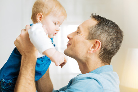 Sweet child. Adorable emotional young father feeling excited while holding his little baby and smiling while trying to kiss himの写真素材