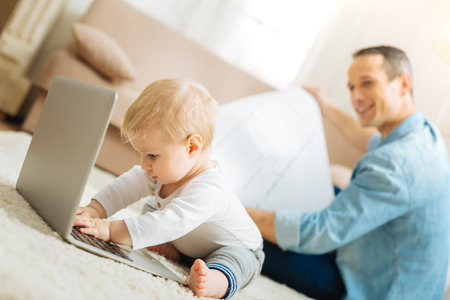 Play time. Lovely little busy baby feeling concentrated while sitting in front of a laptop and trying to type with a kind attentive curious father looking at this funny activityの写真素材