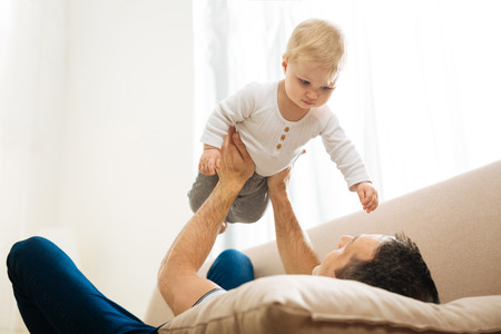 Free time. Enthusiastic caring young father resting on a soft pillow and feeling happy while carrying his lovely little curious childの写真素材