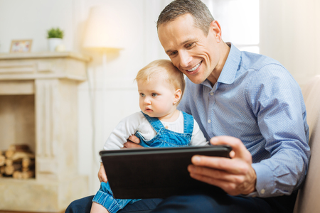 Useful gadget. Cheerful emotional young man feeling excited while holding a baby in his arms and sitting on a comfortable sofa with a modern tabletの写真素材