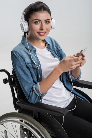 Pleasure in details. Cheerful pretty physically challenged woman putting on headphones while carrying  phone and posing  in the wheelchairの写真素材