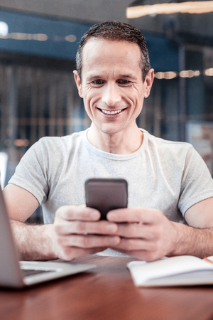 Joyful mood. Positive delighted male person sitting at the table and looking at his gadget while communicating onlineの写真素材