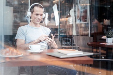 Urban style. Handsome young male sitting in cafe and wearing headphones while using his gadgetの写真素材