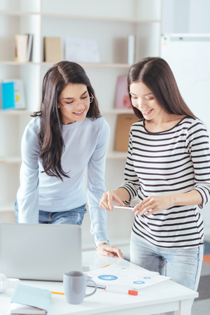 Online cooperation. Joyful merry female colleagues  standing near the table while scanning diagram and preparing reportの写真素材