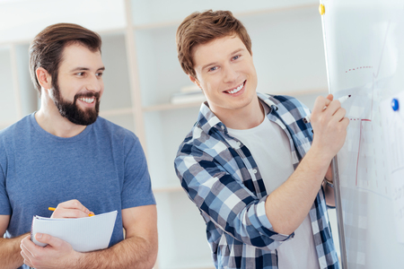 Financial year. Budding  two male colleagues  posing on the blurred background while man smiling near white boar and drawing  at itの写真素材