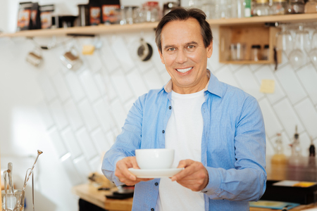 Take it please. Handsome pleasant polite man standing in the kitchen smiling and giving a cup of coffee.の写真素材
