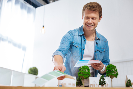Enthusiastic emotional young engineer looking happy while standing in front of miniature houses and smiling when choosing necessary colors from color paletteの写真素材