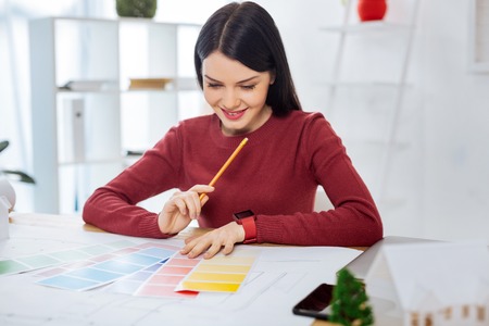 Smart qualified young designer sitting in her modern office with a pencil in one hand and smiling while looking at the color palettes in front of herの写真素材
