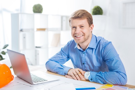 Happy employee. Cheerful smart enthusiastic engineer sitting in front of a modern laptop in his light comfortable office and smiling while feeling happyの写真素材