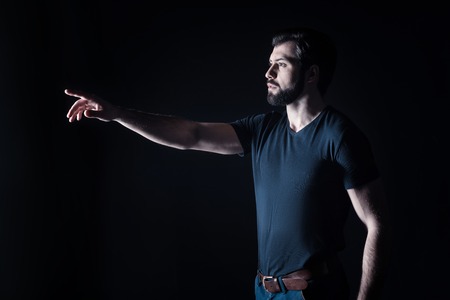 Brutal look. Handsome nice serious man looking in front of him and pointing with his hand while standing against black backgroundの写真素材