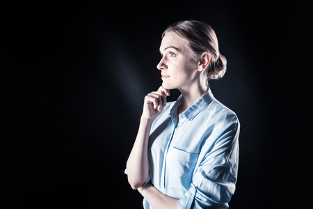 Pleasant thoughts. Nice joyful attractive woman standing against black background and holding her chin while thinkingの写真素材