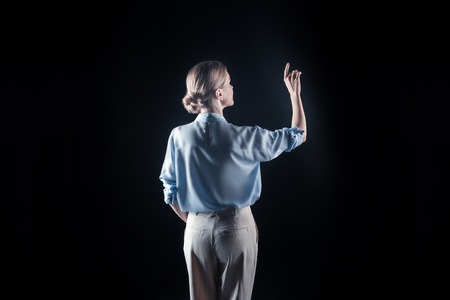 Transparent tech. Pleasant attractive young woman looking in front of her and holding her hand up while touching transparent screenの写真素材