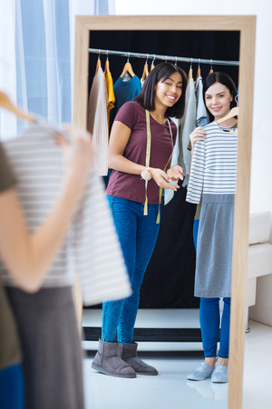 Trying on. Positive friendly young tailor looking happy while standing in front of a mirror and showing a new beautiful dress to a visitor of her atelierの写真素材
