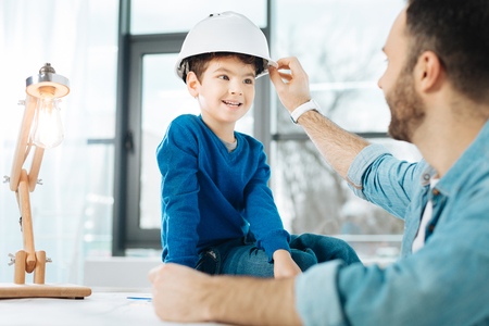 Loving parent. Young loving father fixing a white hard hat on the head of his little son sitting on the top of the table in his office and chatting with himの写真素材