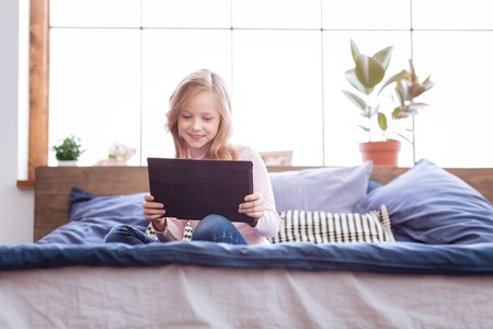 Weekend. Pretty gleeful fair-haired girl smiling and playing games on her tablet while sitting on the bedの写真素材