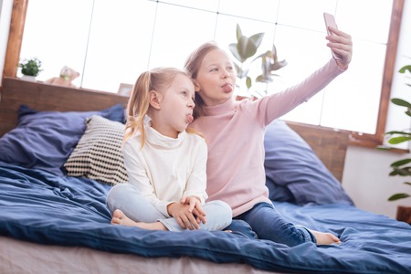 Kidding. Lovely energetic blond sisters smiling and taking pictures on their phone and putting their tongues out while sitting on the bedの写真素材
