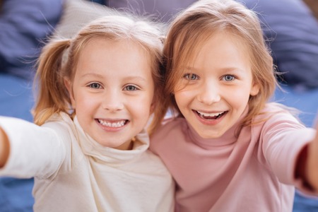 Laughable. Adorable exuberant fair-haired girls smiling and having ponytails and embracingの写真素材