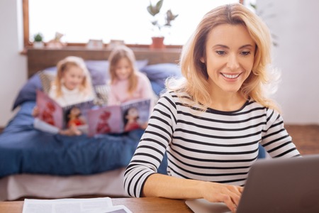 Working. Attractive content blond young mother smiling and working on her laptop while her daughters reading magazinesの写真素材