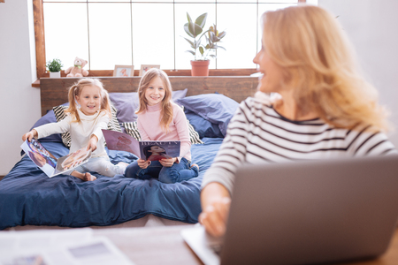 My precious. Cute alert blond little sisters smiling and reading magazines and their mom working and looking at themの写真素材