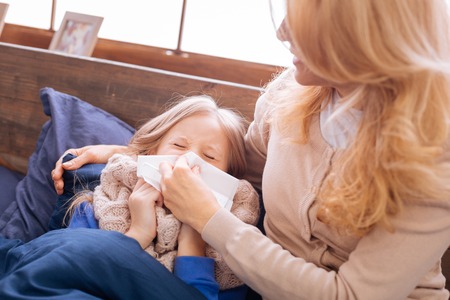 Runny nose. Loving serious blond mother holding a napkin and helping her daughter blow her nose while she lying in bedの写真素材