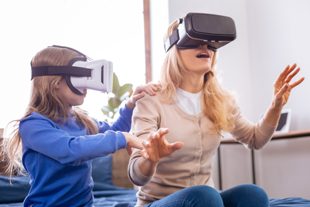 Excited blond mother and daughter wearing VR headset and entertaining while sitting on the bedの写真素材
