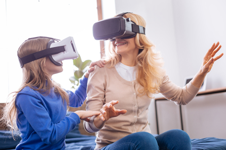 Cheerful fair-haired young mother and daughter wearing VR headset and entertaining while sitting on the bedの写真素材