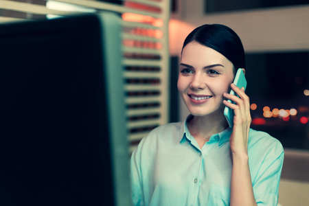 Pleasant conversation. Upbeat pretty young woman talking on the phone while working on the computer in the office and smilingの写真素材