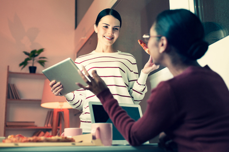 Non-formal ambience. Two pleasant female colleagues eating pizza and talking to each other, discussing work, while one of them holding a tabletの写真素材