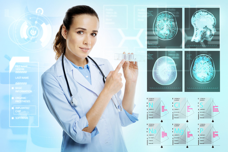 Useful box. Young professional calm doctor standing near the scull xray with a stethoscope on her neck and pointing to a tiny pillbox in her handsの写真素材