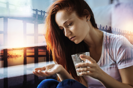 Young tired ill woman feeling unwell and looking at the pill in her hand while holding a glass of waterの写真素材