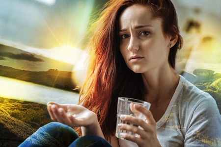 Tired ill young woman looking upset while sitting with a glass of water and holding necessary pills in her handの写真素材