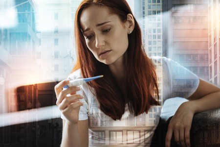 Ill upset disappointed woman sitting on the sofa looking at the thermometer and feeling bad herself.