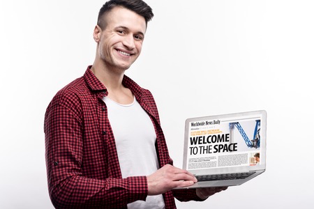Being all-rounder. Handsome pleasant young man in a checked shirt showing a laptop with a news...