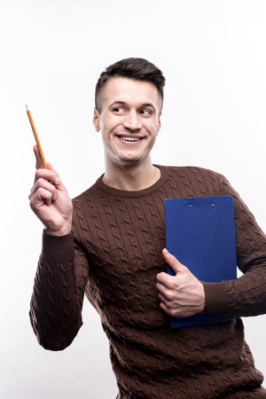 Upbeat office worker. Cheerful young man in a brown sweater posing with a pencil and a blue sheet holder while smiling and standing isolated on a white backgroundの写真素材
