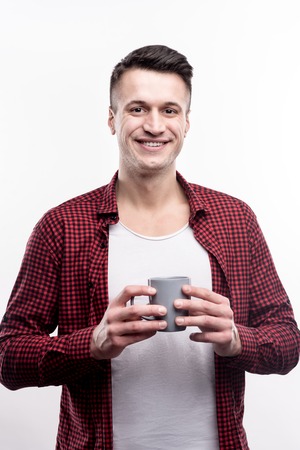 Healthy drink. Charming young man in a checked shirt holding a cup of tea with his both hands while posing against a white backgroundの写真素材