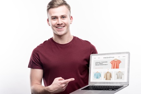My best project. Handsome fair-haired young man in a burgundy t-shirt pointing at a laptop with...