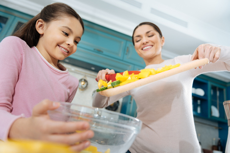 Cooking process. Pretty inspired dark-haired mother smiling and putting ingredients into a bowl and making a salad with her daughterの写真素材
