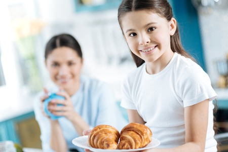 Help yourself. Pretty happy dark-eyed little girl smiling and holding a plate with some croissants and her mom sitting in the backgroundの写真素材
