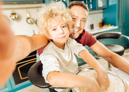 Special recipe. Cute smart skillful boy standing together with his father and putting hands into the bowl with flour while preparing doughの写真素材