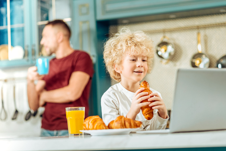 Delicious snack. Nice positive happy boy smiling and looking at the laptop screen while eating a croissantの写真素材