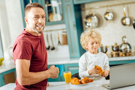 At home. Nice pleasant man holding a cup of tea and looking at you while being with his son in the kitchenの写真素材