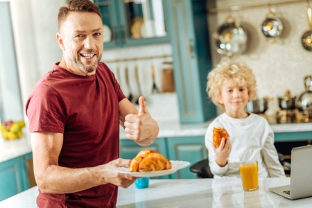 Good job. Happy positive delighted man standing in the kitchen and looking at you while holding a plate with croissantsの写真素材